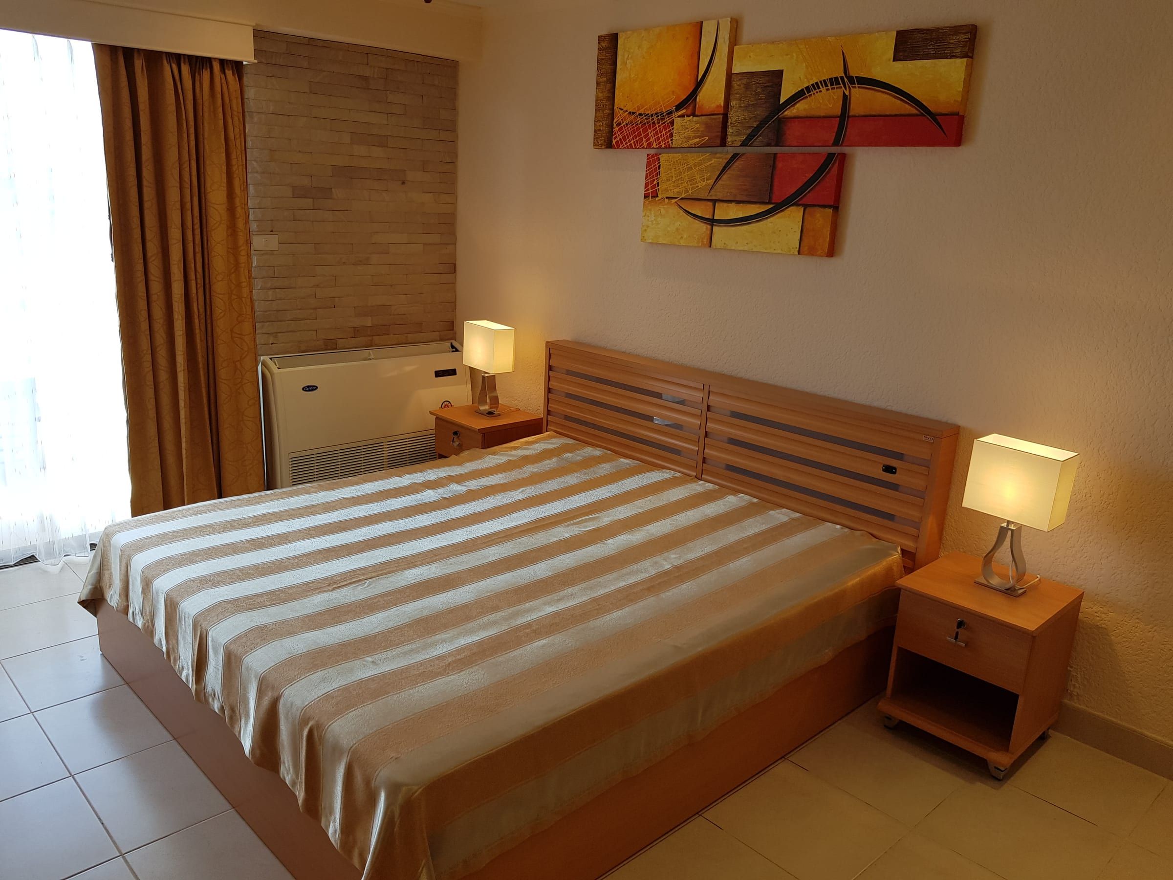 Studio Typ A: Kingsize-Bett mit gestreifter Tagesdecke, hellem Holzkopfteil, Nachttischlampen und Bildern an der Wand