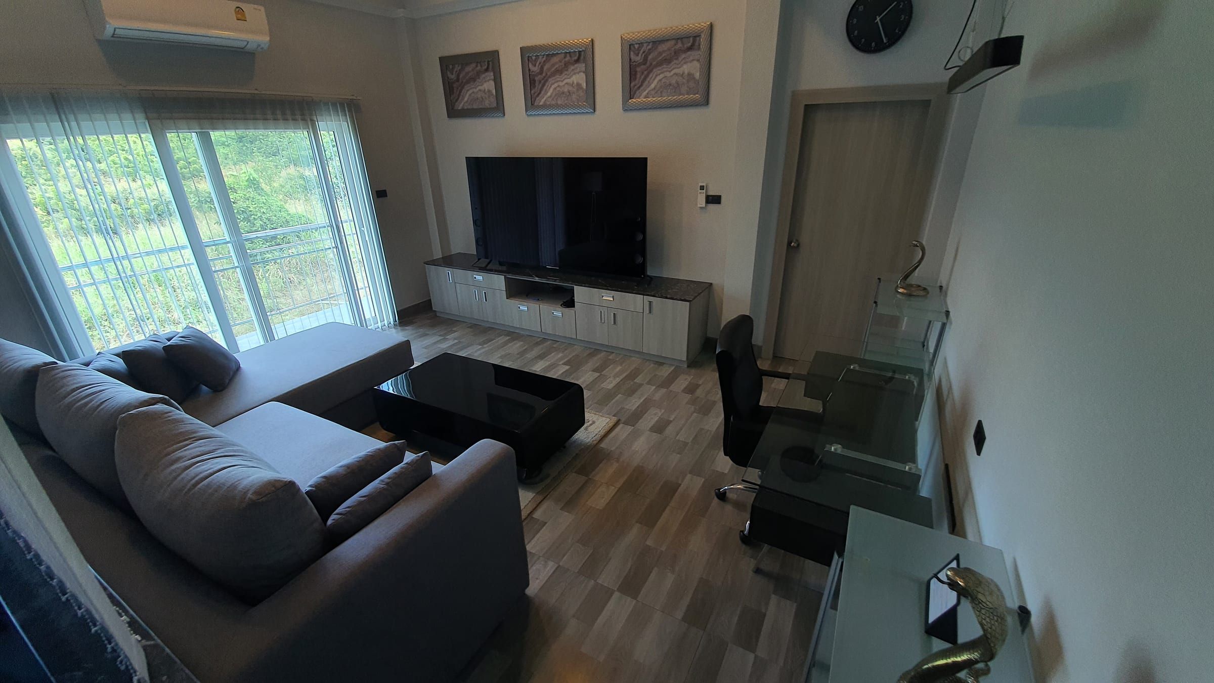Wohnbereich der Loft-Wohnung mit grauem Sofa, großem Fernseher, Schreibtisch und Fenster ins Grüne