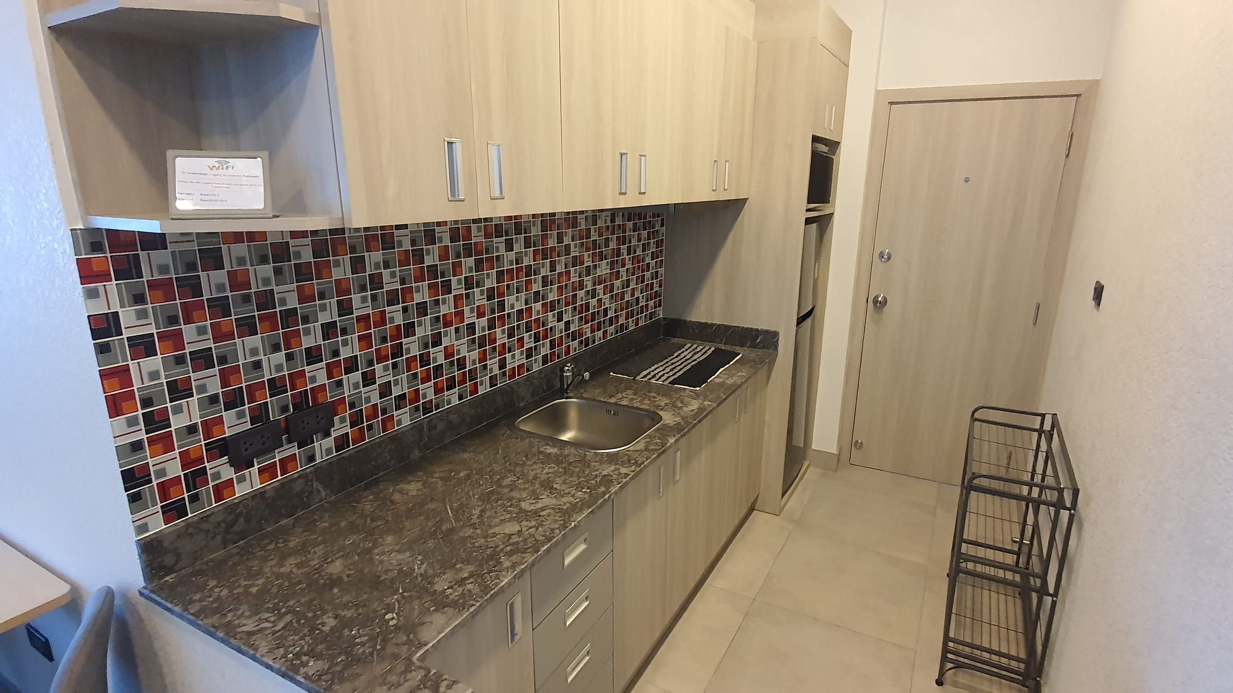 Kitchenette avec placards en bois clair, plan de travail en pierre sombre, évier et mosaïque rouge, noire et blanche, porte d'entrée au fond