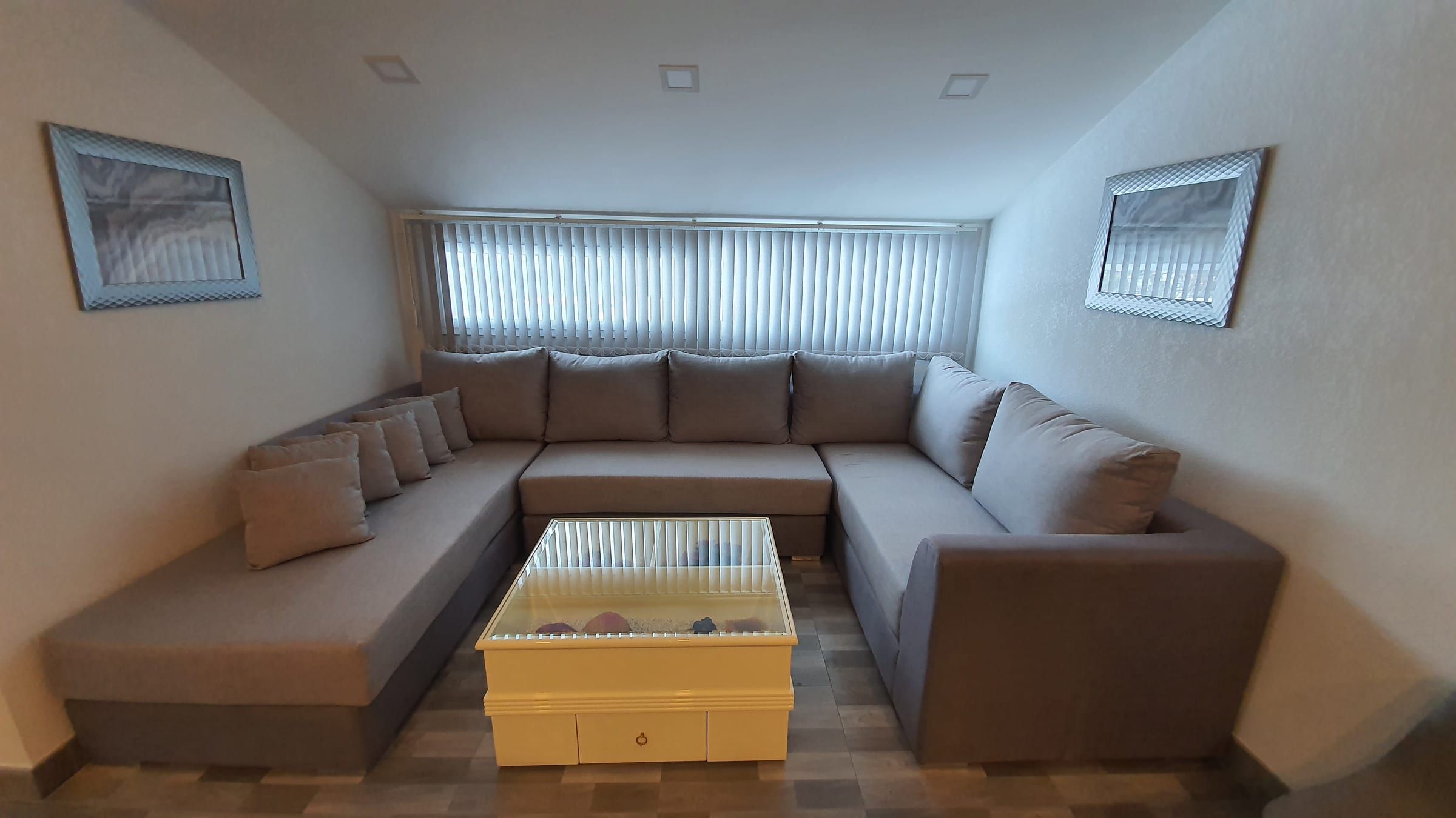 Espace de vie avec grand canapé gris en U et table basse blanche à plateau de verre sous le plafond mansardé