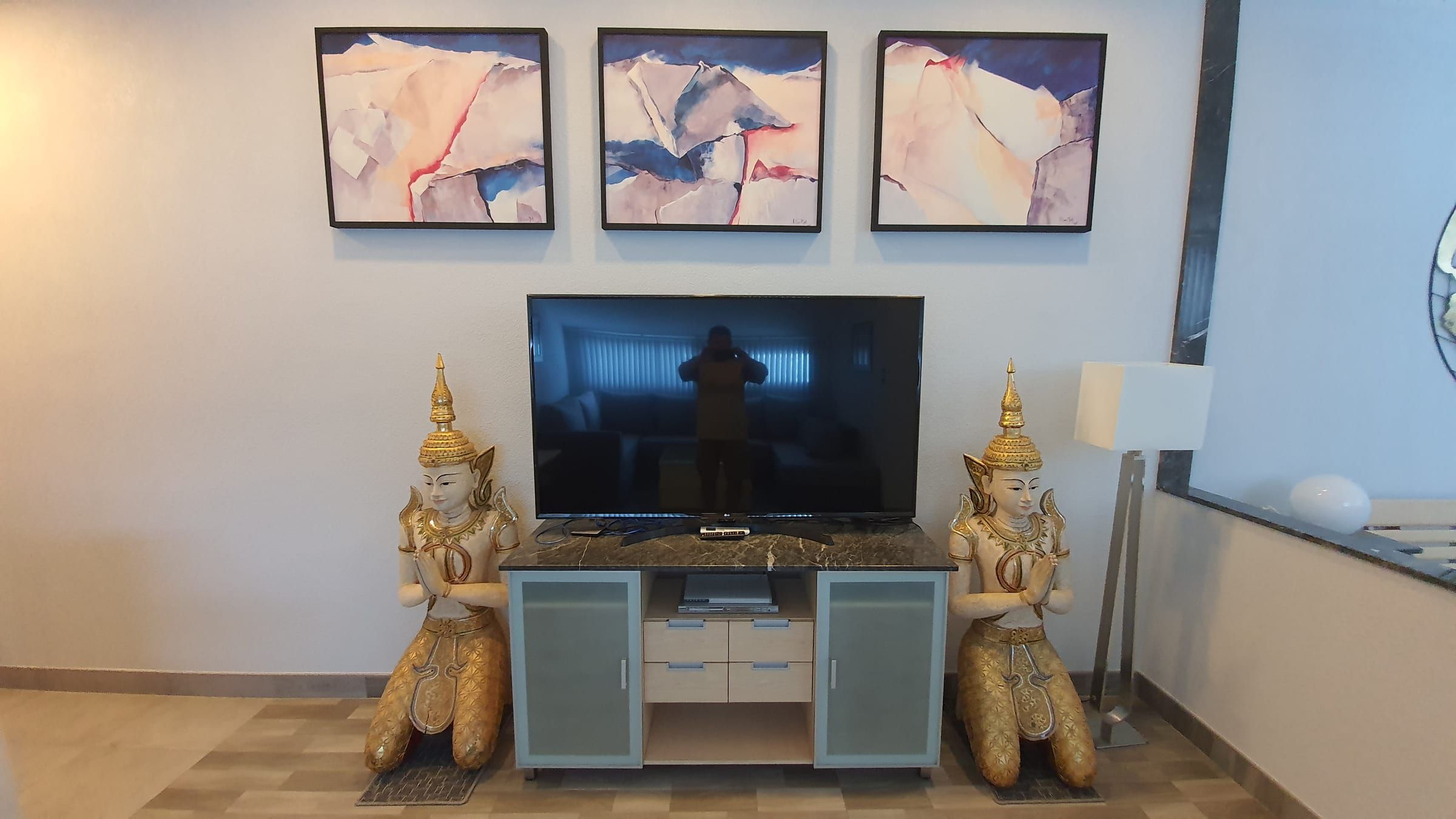 Téléviseur sur un buffet entre deux statues thaïes dorées en position de prière, surmonté de trois tableaux abstraits